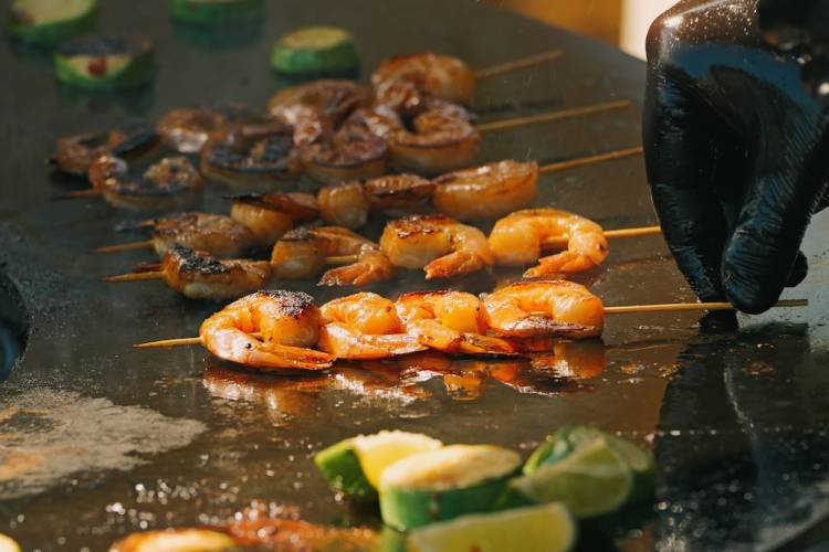 Close-up of a chef cooking seasoned shrimp skewers on a sizzling hot flat-top grill.